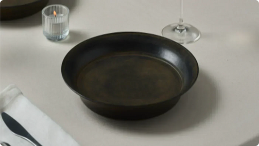A dark brown rimmed bowl on an off-white table, a ribbed glass candle and a wine glass beside it, linen napkin at the edge