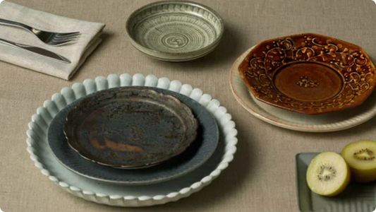 Plates laid on linen — blue-grey and rust-toned plates stacked on a white petal-rimmed dish, a ridged ash-glazed bowl, an octagonal amber-glazed plate with chatter marks