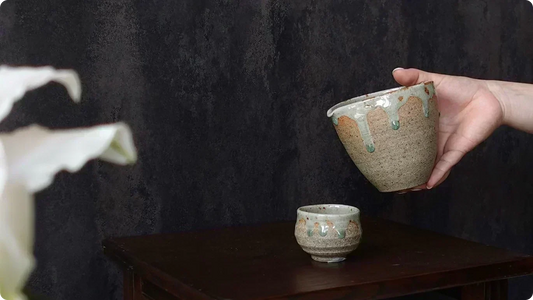 Against a black wall, a hand holds a white-slip katakuchi above a small guinomi on a wooden stand, green-tinged glaze running from the rim like droplets
