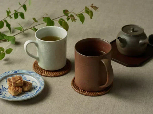 A white porcelain mug and a red clay mug on woven rattan coasters over linen, with a blue-and-white plate of baked sweets at left and a red clay teapot behind