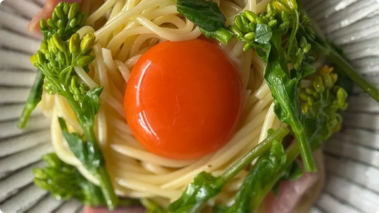 Close-up of a white ridged plate holding a nest of pasta with rapeseed blossoms and ham, an orange yolk resting at the centre