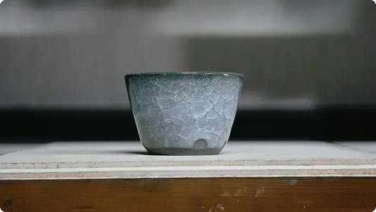 A blue-grey tea cup on a board, white crazing running across its whole surface and a deeper green gathering only at the rim
