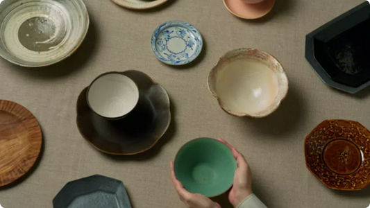 Ware from across Japan scattered on linen, seen from above — an ash-glazed bowl, a blue-and-white small plate, a black petal-rimmed dish, a white slip bowl, an amber octagonal plate, and a celadon bowl held in two hands