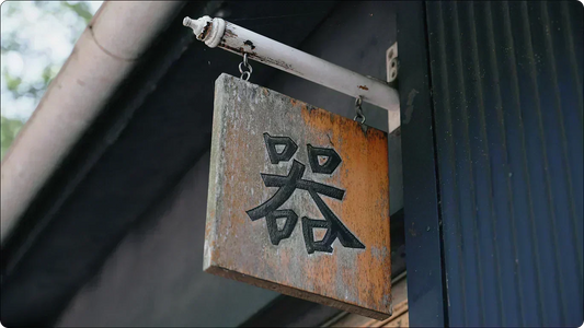 A weathered wooden shop sign carved with the character for utsuwa, rust and moss marking its face beneath the eaves