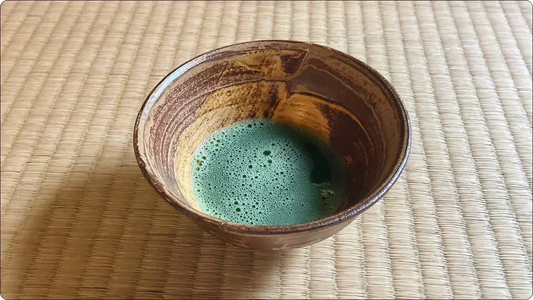 畳の上に置かれた茶碗に点てられた抹茶。無釉の器肌に炎が残した緋色の景色が流れる