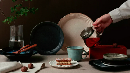 Ware on a dim table — a ridged black plate and a large unbleached plate propped up, a blue-green mug, a mille-feuille on a petal-rimmed dish, tea poured from a glass pot onto a red lacquer box
