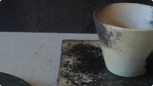 An unglazed cylindrical cup and square plate, ash-darkened black patches spreading across the pale, bare clay surface