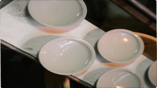 薄暗い工房で、板（乾燥用のトチ／敷き板）の上に伏せて並ぶ、艶のある淡い白釉の粉引の浅皿