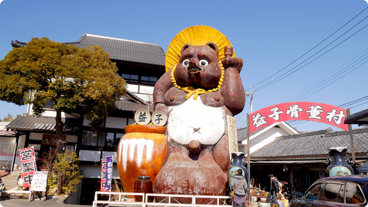 A giant ceramic tanuki standing on a street corner in Mashiko, wearing a straw hat and raising a sake flask, a large amber-glazed jar at its feet and a red antique-village sign behind