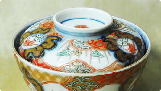 Close-up of an overglaze-enamelled lidded bowl, gold outlining the cloud shapes on the lid and a gold lattice on the body over white porcelain and red