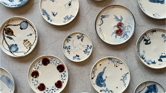 Hand-painted plates by Higuchi Maki laid out on stone: lanterns, a crane with persimmons, rabbits, pandas and an elephant in cobalt and iron red