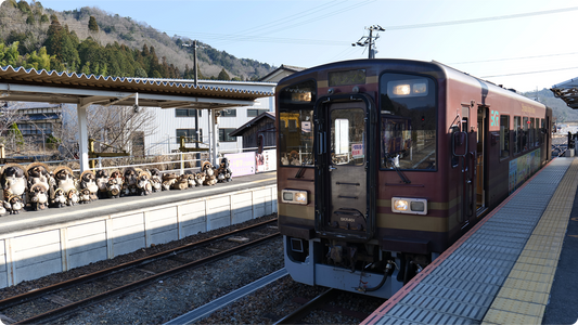 信楽駅のホームに停まる信楽高原鐵道の1両編成。向かいのホームぎわには笠をかぶった信楽たぬきの置物がずらりと並び、背後に山が見える