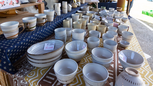 A stall at the Folk Pottery Village Festival: white cups, bowls and oval dishes banded with fine tobikanna chatter marks, price tags beside them, on indigo-dyed cloth