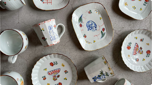 Fluted porcelain plates and mugs by TATA pottery studio laid out on stone, hand-painted with a blue rabbit, a sleeping cat and scattered red and yellow flowers