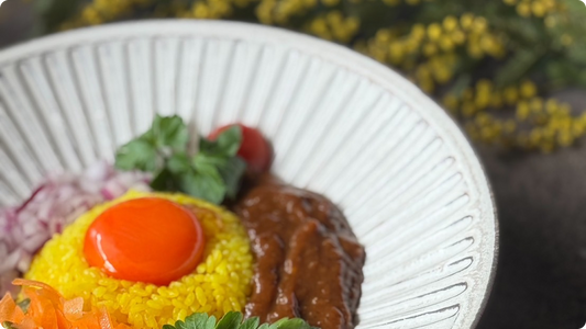 A white rimmed plate carved with radiating ridges, holding turmeric rice with an egg yolk, curry, red onion and a cherry tomato, with mimosa blurred behind