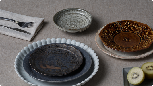 Plates laid on linen — a black plate stacked on a pale blue foliate rimmed plate, a gray dish carved with a spiral, and an amber foliate plate. Cutlery to the left, kiwi at lower right