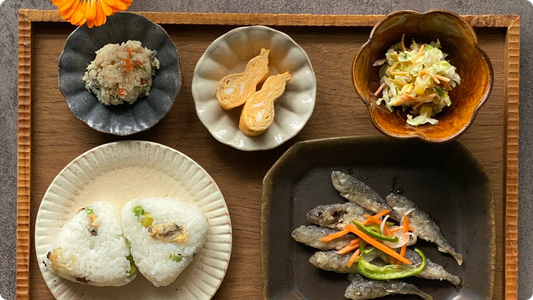 A wooden tray set with small plates — seasoned rice on a black foliate mamezara, inari sushi on a white foliate dish, a vinegared salad in a brown bowl, two onigiri on a foliate plate and small fried fish on a black square dish