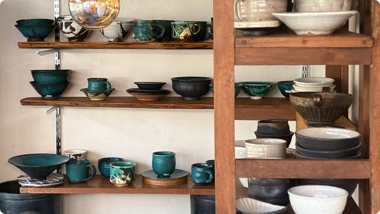 Pottery on wooden shelves — bowls and cups in a turquoise glaze alongside black bowls and saucers, with white and black pieces stacked on the shelves to the right