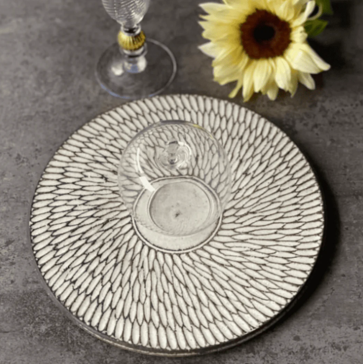 A round white platter carved with fine radiating strokes, set on gray stone with a glass and a sunflower