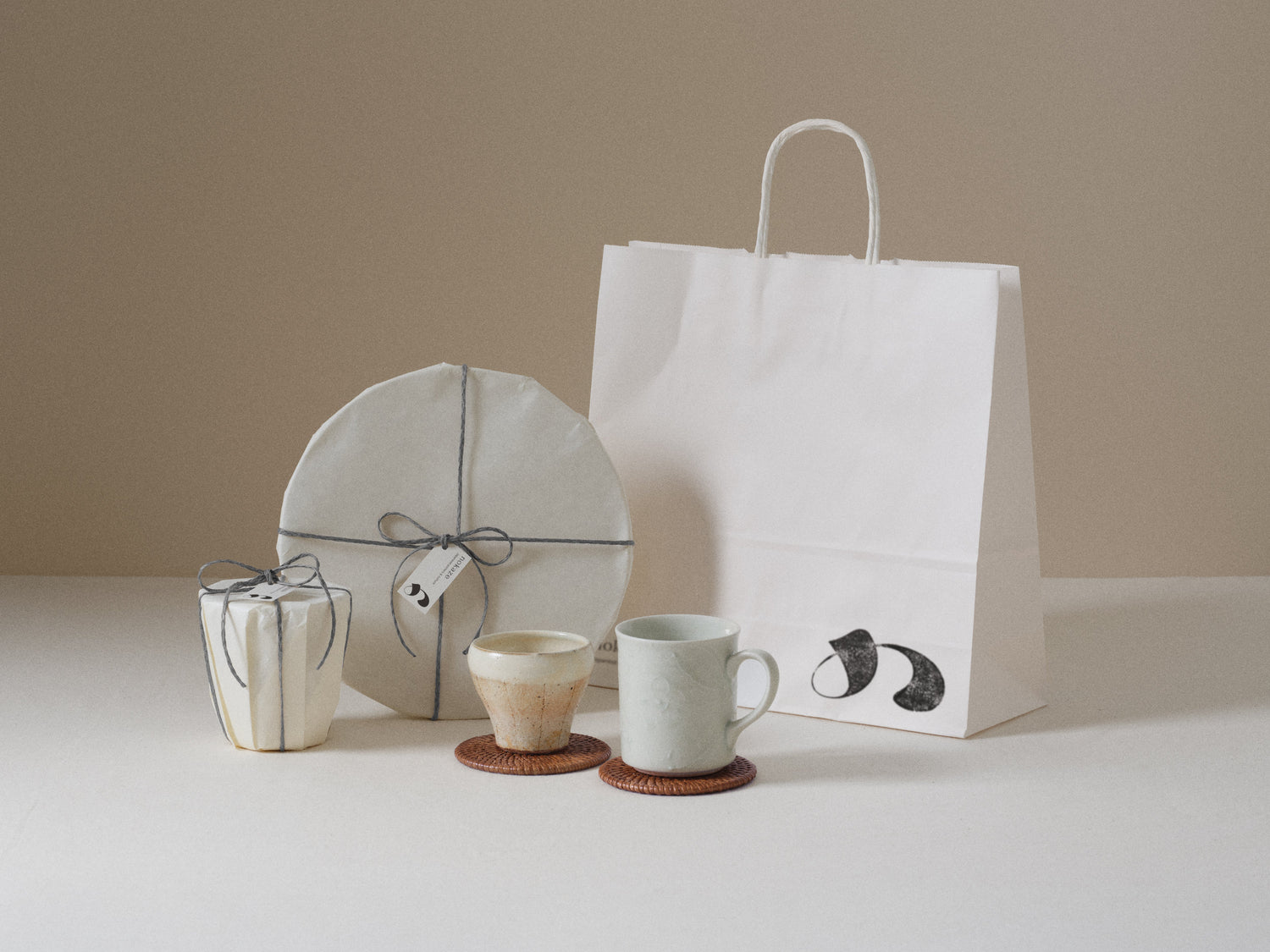 Two pieces wrapped in white paper and tied with grey cord, tagged with the Nokaze mark, beside a cup, a mug and a paper bag