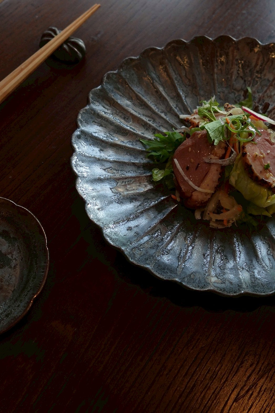 A large iron glazed chrysanthemum plate of seared duck salad, with chopsticks on a rest on dark wood