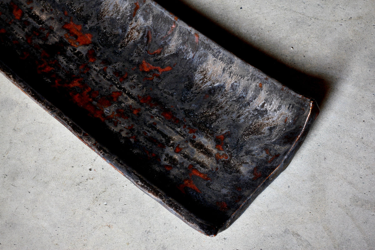 A long black glazed rectangular plate streaked with rust orange, resting on a pale concrete floor