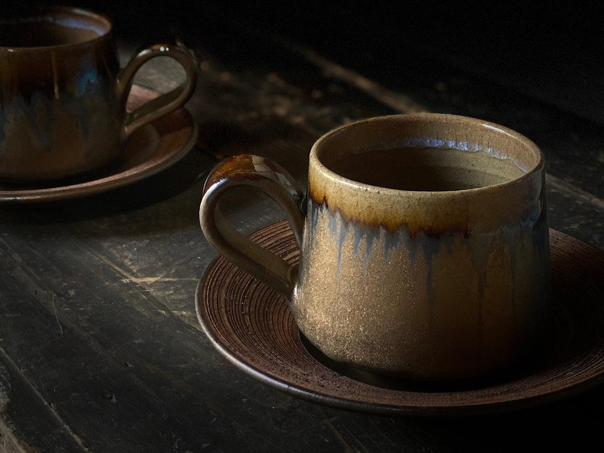 Two brown glazed cups on matching saucers, their amber rims running into pale blue drips, on dark wood