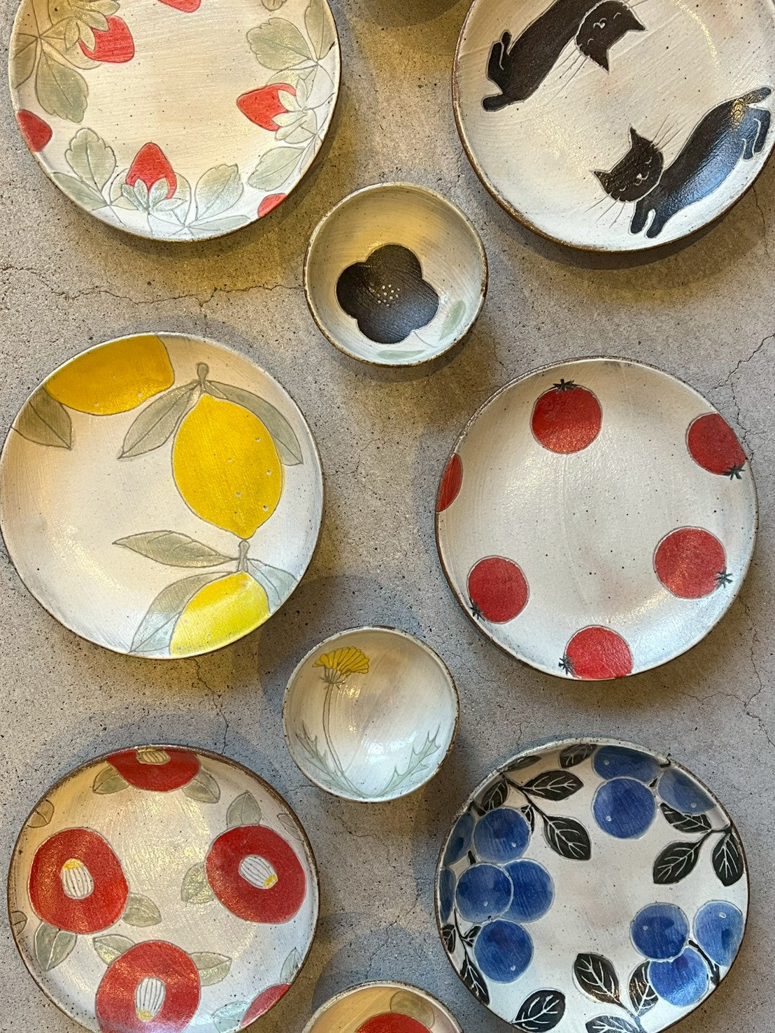Hand painted plates and small bowls scattered on concrete — lemons, tomatoes, camellias, berries and cats