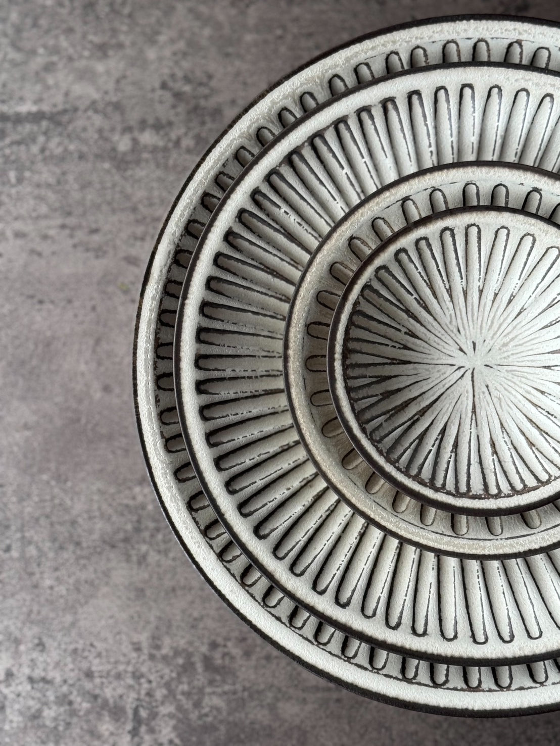 Stacked white plates carved with radiating shinogi facets, their dark rims showing, on gray concrete