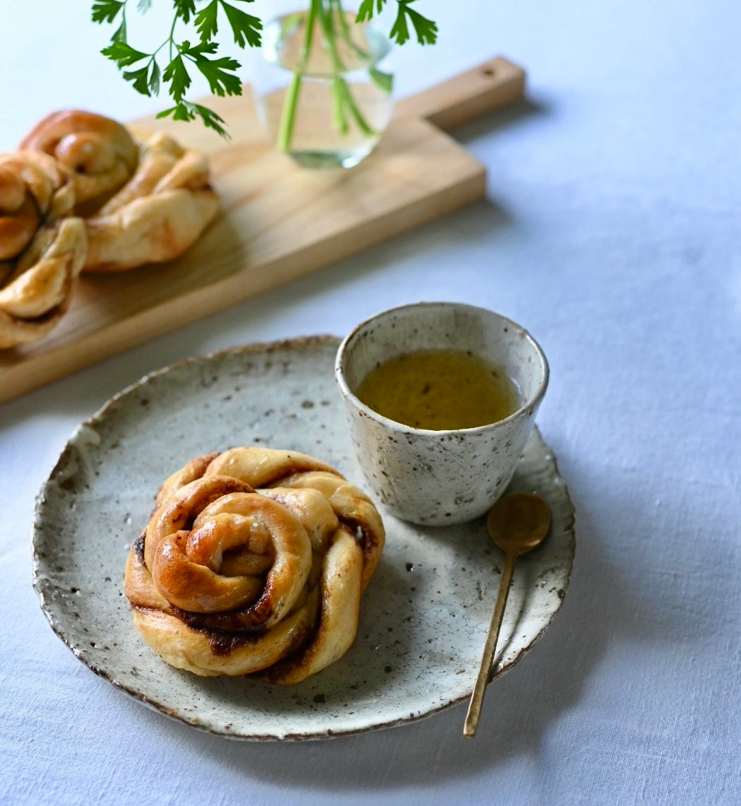 A speckled white medium plate holding a cinnamon bun, with a matching cup of tea and a brass spoon